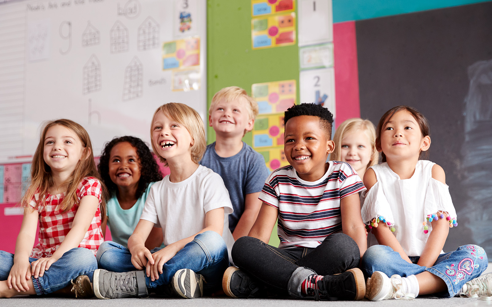 A group of children sitting on the floor in a classroom setting, smiling and looking towards the camera.