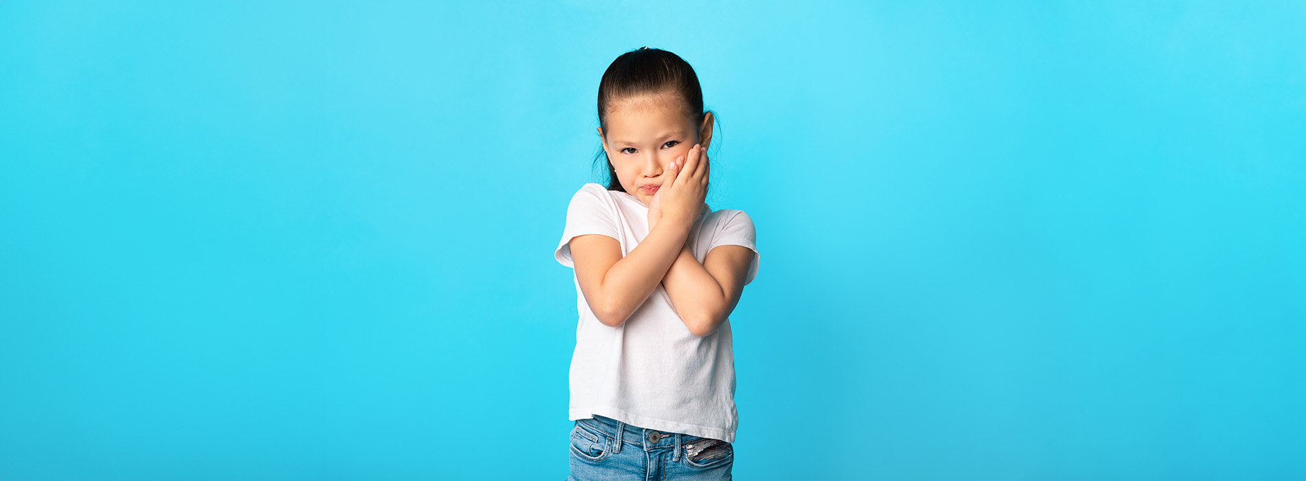 A young child standing against a blue background, holding their hands to their cheeks.