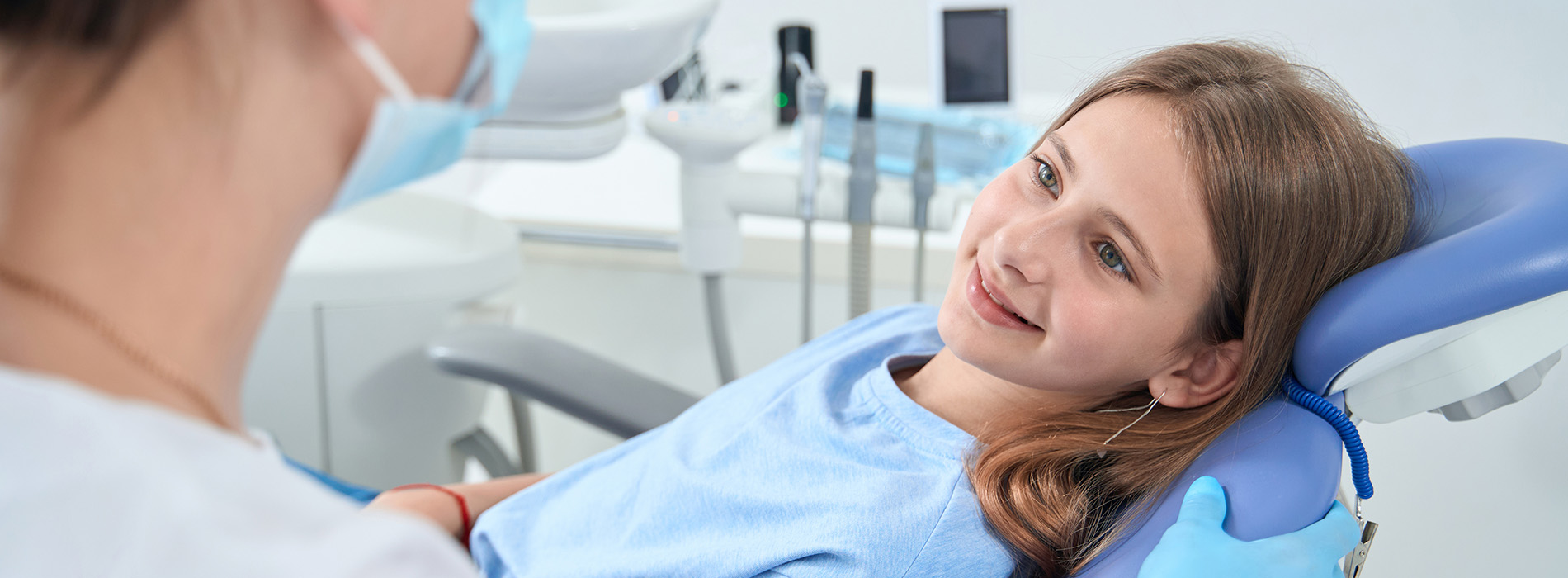 A young woman is seated in a dental chair, smiling at the camera, while a female dentist attends to her.