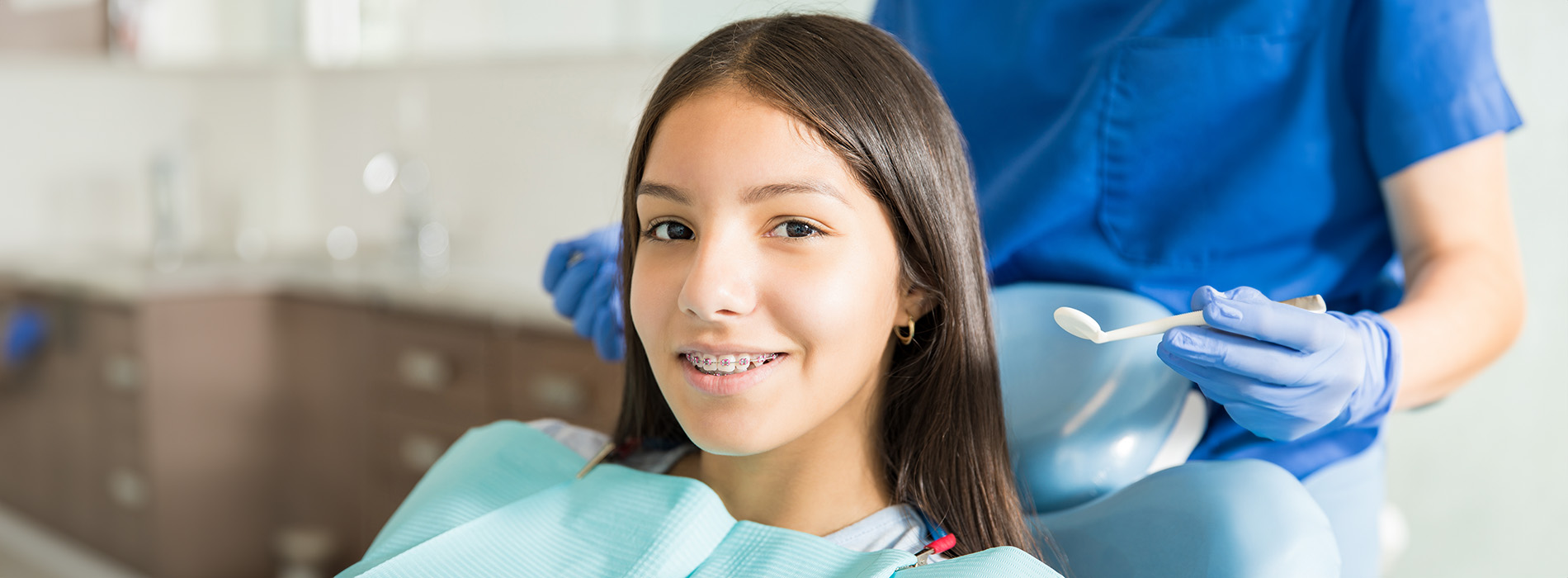 The image shows a young woman sitting in a dental chair, receiving dental care from a professional wearing blue gloves and a white coat.