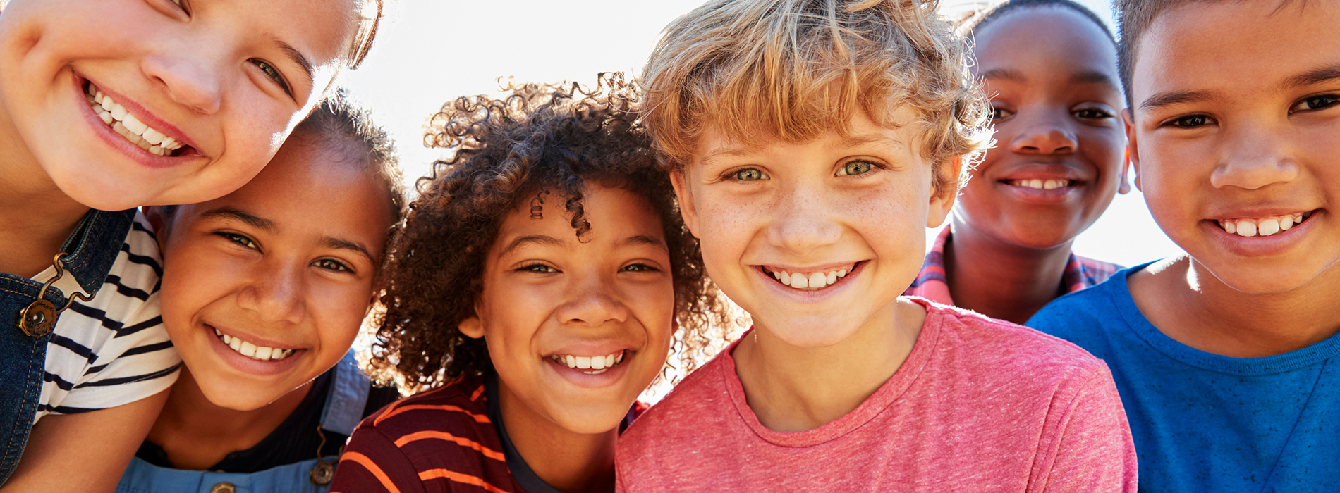 The image shows a group of children smiling at the camera, with one child appearing to be looking away from the group. They are outdoors during the daytime.