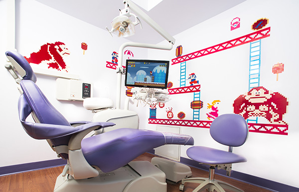 The image shows a dental chair with a purple seat and headrest, situated in a room decorated with colorful wallpaper featuring cartoon characters and a gaming theme.