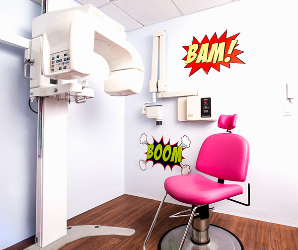 The image shows an interior space with a medical examination room setting, featuring a large, modern imaging machine, a pink chair with a comic-style sticker on the wall behind it that says  BOOM  with an explosion graphic, and a playful, cartoonish sticker above the chair with text that reads  BAM   and a graphic of a bomb. The room appears to be well-lit and clean, with medical equipment and a blue color scheme.